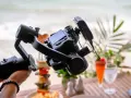 Un gimbal stabilizza una fotocamera per riprese in stile mezzo busto cinema, con un drink e cibo in spiaggia sullo sfondo.