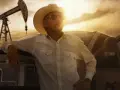 Uomo con cappello da cowboy e camicia a righe appoggiato a un pick-up, con pozzi petroliferi sullo sfondo. Il western significato è qui, tra lavoro e orizzonti infiniti.