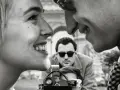 Primo piano di una coppia che ride, con un uomo che filma sullo sfondo, evocando l'estetica dei registi della nouvelle vague.