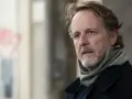 Christian Peppard, con un foulard e un cappotto scuro, guarda pensieroso verso destra.