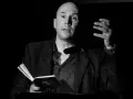 Un uomo calvo, narratore protagonista, legge da un libro aperto, gesticolando con la mano.