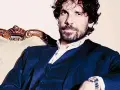 L'attore Montanari, con barba e capelli ricci, indossa un completo blu e una camicia bianca, seduto su una poltrona d'epoca.
