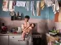 Emiliano Dante, padre single, nutre il suo bambino in una cucina disordinata, con panni stesi ad asciugare.