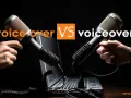 Due microfoni professionali, uno tenuto da una mano, l'altro da un'altra, si fronteggiano sopra un laptop. Il testo "voice over VS voiceover" suggerisce una competizione o un confronto nel mondo del doppiaggio e della narrazione.