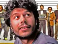 Un primo piano del protagonista di un film di Sydney Sibilia, con altri personaggi sullo sfondo che tengono cartelli con scritte come "CHIMICO" e "NEURO BIOLOGO".