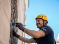 Uomo con casco giallo dipinge una grata metallica con un pennello blu. Inquadratura dal basso, cielo azzurro sullo sfondo.