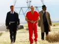 Tre uomini camminano in un campo: un detenuto in tuta rossa ammanettato, un poliziotto e un detective. Scena da film polizieschi recenti.