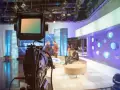 Telecamera in primo piano in uno studio televisivo. Si intravede il backstage, con persone sedute e in piedi, luci soffuse e un'atmosfera di attesa.