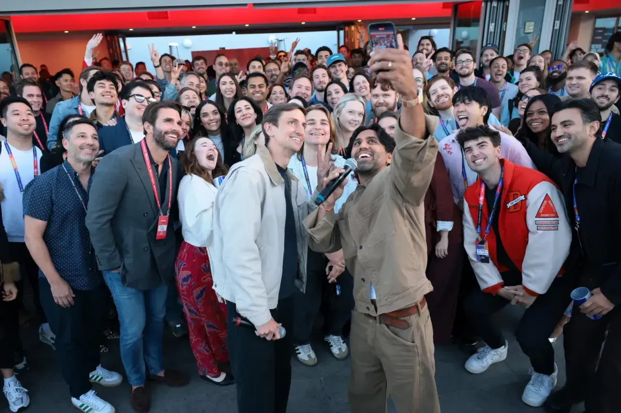 Un gruppo di persone sorride e fa un selfie a Hollywood, America. Alcuni tengono microfoni, creando un'atmosfera di festa.
