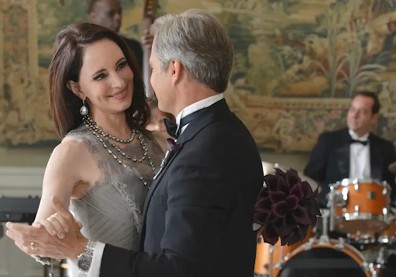 Il cast di Revenge balla elegantemente. Una donna con un abito grigio e collane di perle, un uomo in smoking.