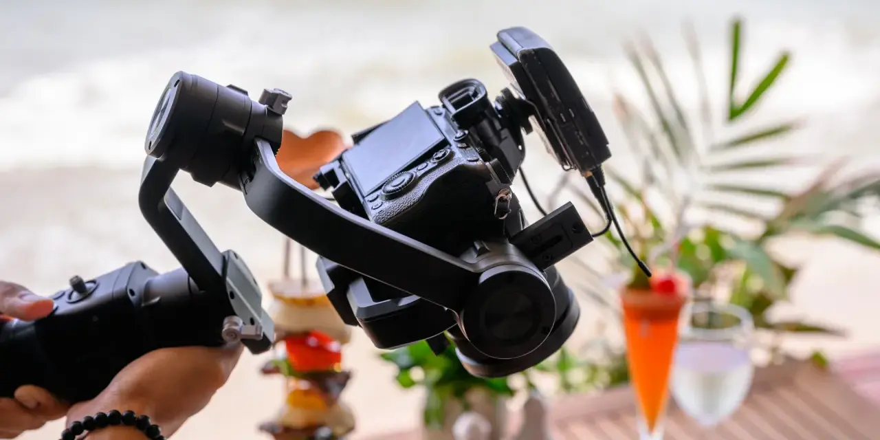 Un gimbal stabilizza una fotocamera per riprese in stile mezzo busto cinema, con un drink e cibo in spiaggia sullo sfondo.