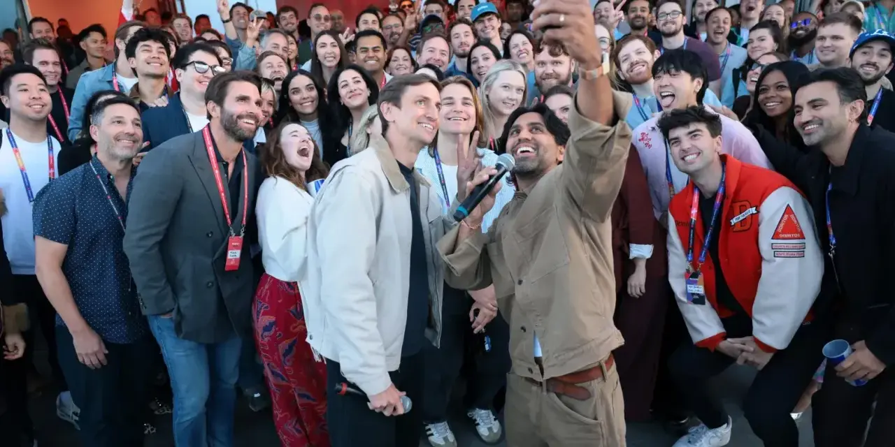 Un gruppo di persone sorride e fa un selfie a Hollywood, America. Alcuni tengono microfoni, creando un'atmosfera di festa.
