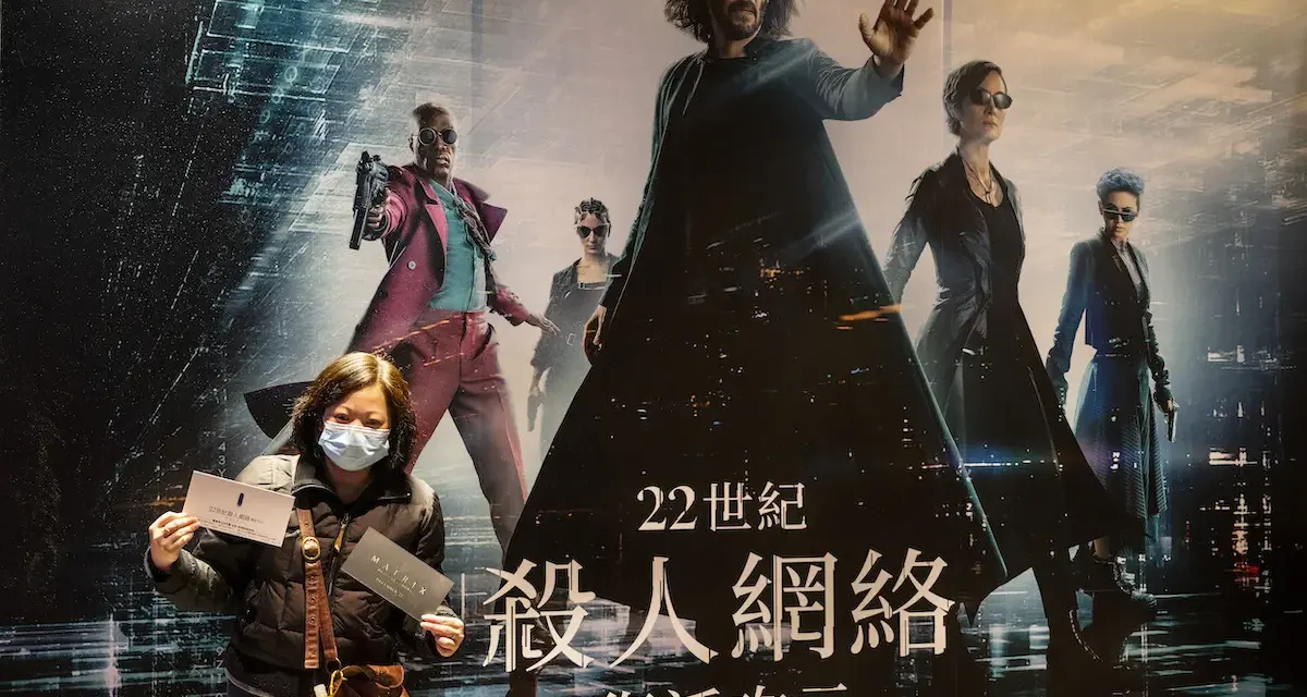 Donna con mascherina mostra biglietti davanti al poster di Matrix, con Neo e Trinity in primo piano. La trama di Matrix è avvincente.