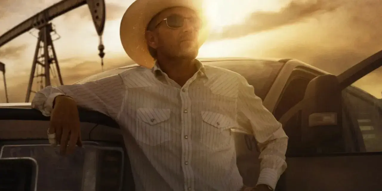 Uomo con cappello da cowboy e camicia a righe appoggiato a un pick-up, con pozzi petroliferi sullo sfondo. Il western significato è qui, tra lavoro e orizzonti infiniti.