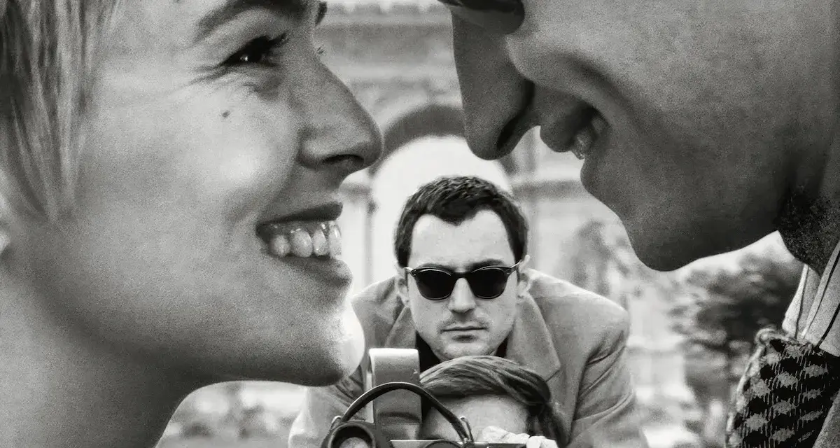 Primo piano di una coppia che ride, con un uomo che filma sullo sfondo, evocando l'estetica dei registi della nouvelle vague.
