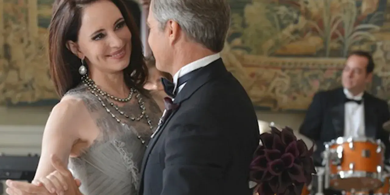Il cast di Revenge balla elegantemente. Una donna con un abito grigio e collane di perle, un uomo in smoking.
