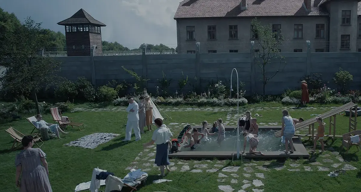 Scena da film strani: bambini giocano in piscina, adulti sorvegliano, mentre sullo sfondo incombono torri di guardia e un edificio imponente.