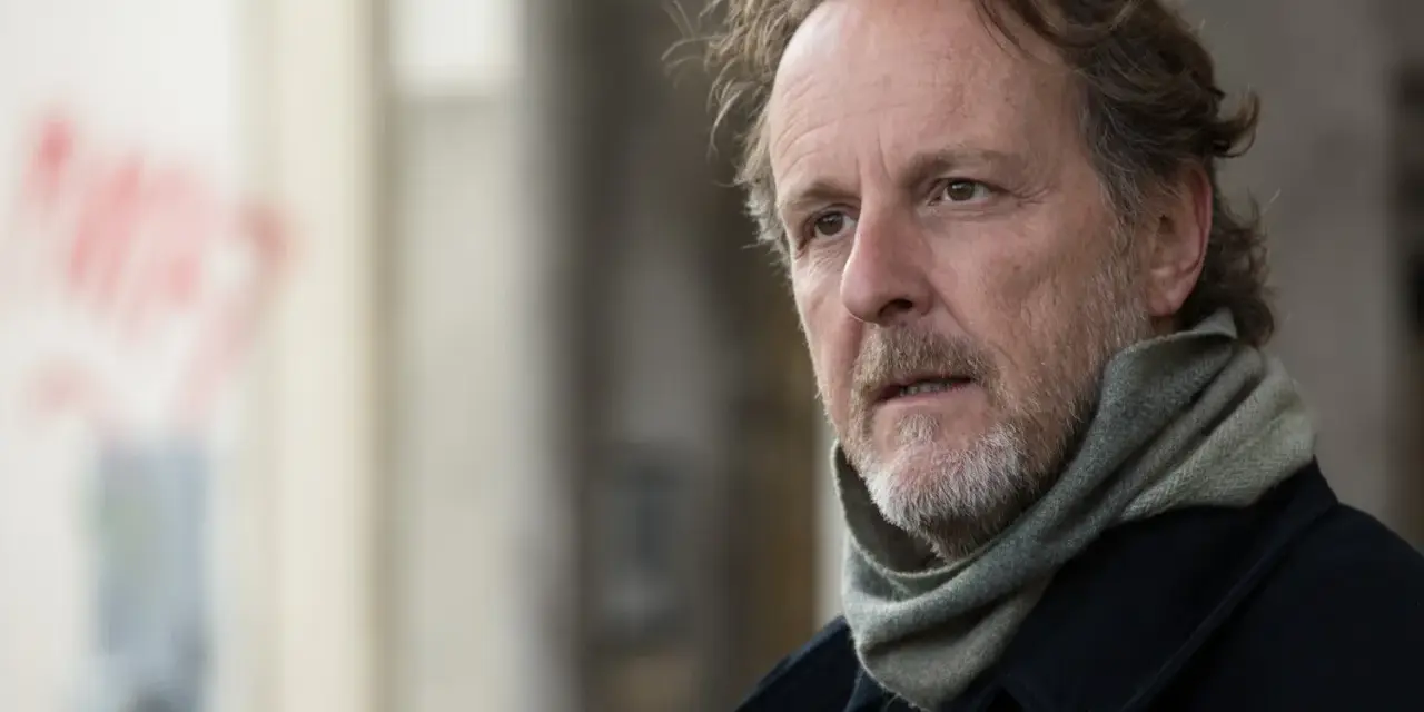 Christian Peppard, con un foulard e un cappotto scuro, guarda pensieroso verso destra.