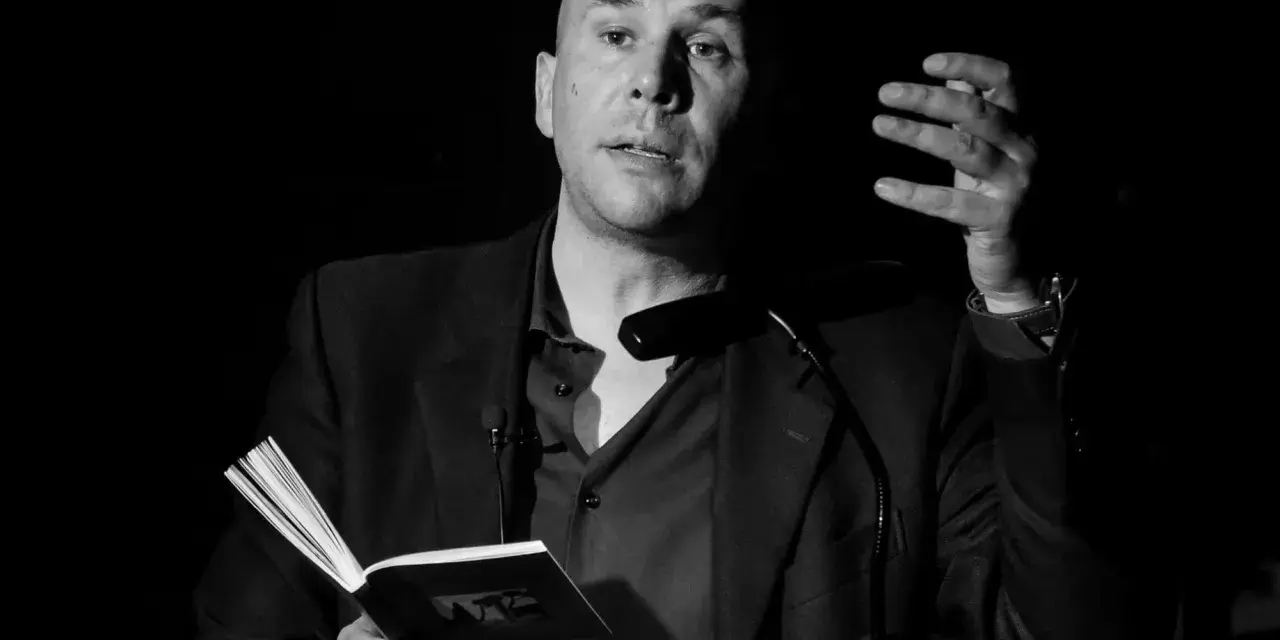 Un uomo calvo, narratore protagonista, legge da un libro aperto, gesticolando con la mano.