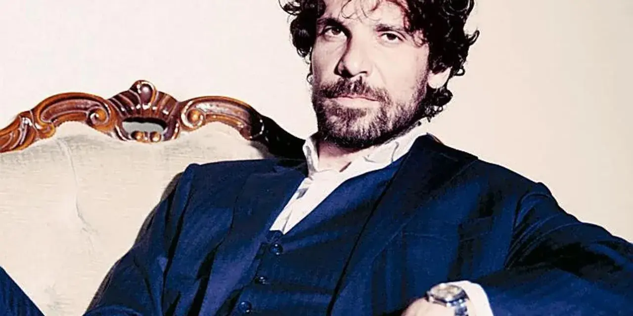 L'attore Montanari, con barba e capelli ricci, indossa un completo blu e una camicia bianca, seduto su una poltrona d'epoca.