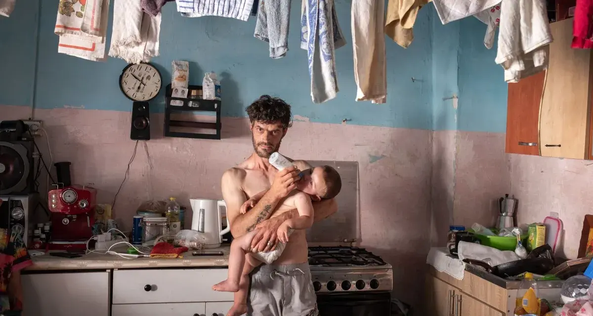 Emiliano Dante, padre single, nutre il suo bambino in una cucina disordinata, con panni stesi ad asciugare.
