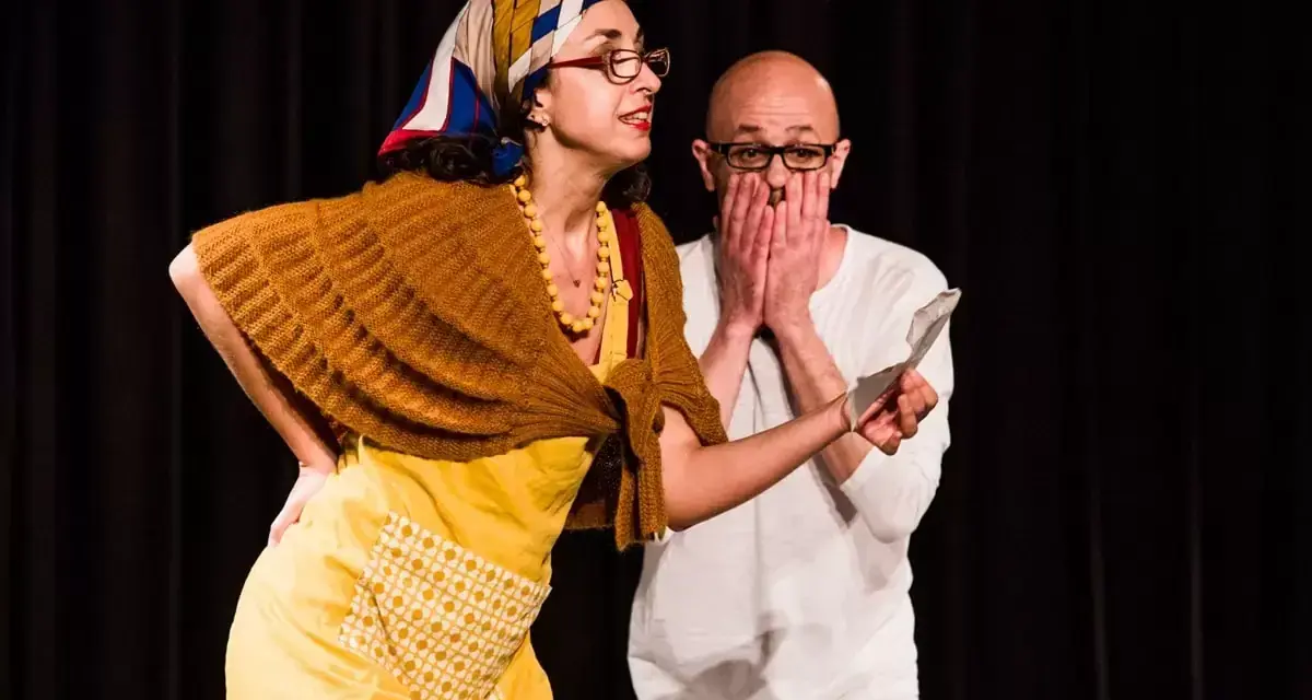 Due attori in scena, una donna con foulard e grembiule giallo, un uomo con le mani sul volto. Una scena di commedia drammatica.