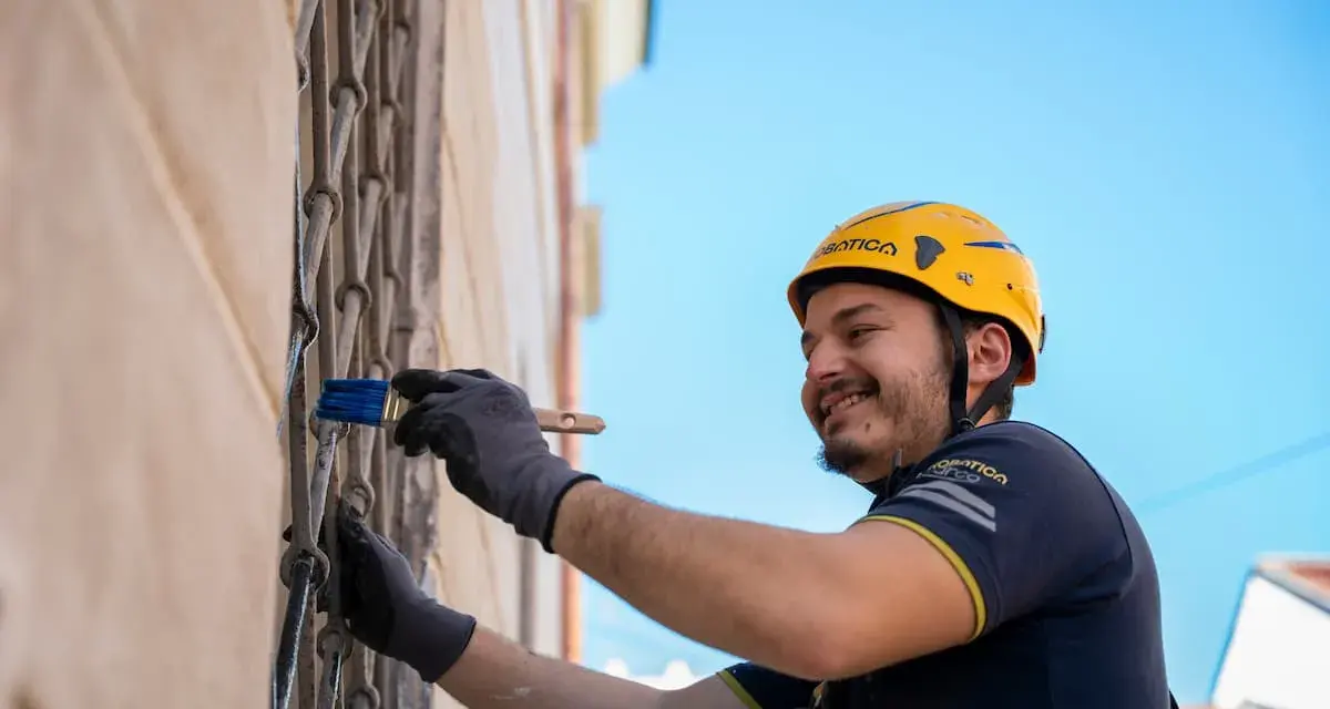Uomo con casco giallo dipinge una grata metallica con un pennello blu. Inquadratura dal basso, cielo azzurro sullo sfondo.