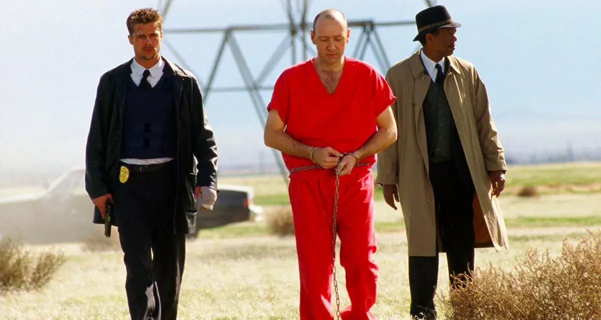 Tre uomini camminano in un campo: un detenuto in tuta rossa ammanettato, un poliziotto e un detective. Scena da film polizieschi recenti.