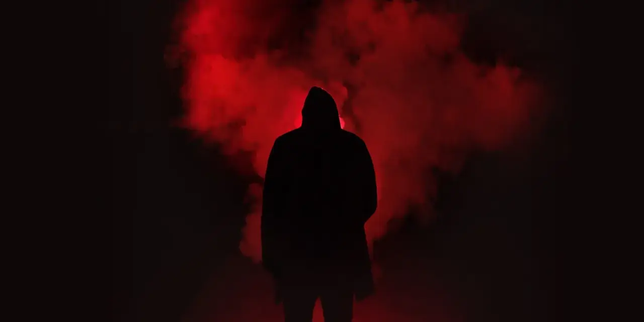 Figura incappucciata nell'oscurità, circondata da fumo rosso. Un'immagine che evoca la differenza tra giallo e noir, tra mistero e oscurità.
