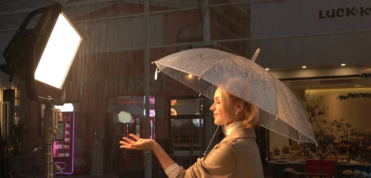 Una donna con ombrello trasparente sotto la pioggia, illuminata da potenti luci set cinematografico. La scena evoca un'atmosfera suggestiva e cinematografica.