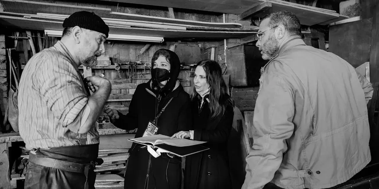 Foto di scena: un artigiano discute con due donne, una con mascherina, mentre un uomo osserva.