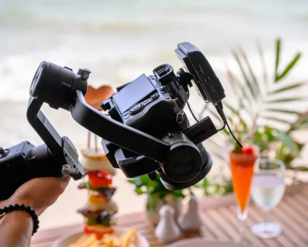 Un gimbal stabilizza una fotocamera per riprese in stile mezzo busto cinema, con un drink e cibo in spiaggia sullo sfondo.