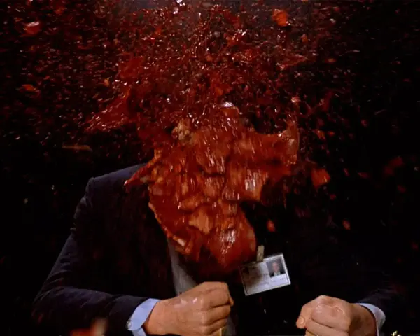 Un uomo in giacca e cravatta, vittima di un evento cruento, con sangue e detriti che esplodono in un'immagine tipica di un genere cinematografico con scene truculente.