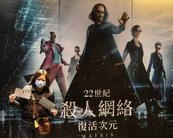 Donna con mascherina mostra biglietti davanti al poster di Matrix, con Neo e Trinity in primo piano. La trama di Matrix è avvincente.
