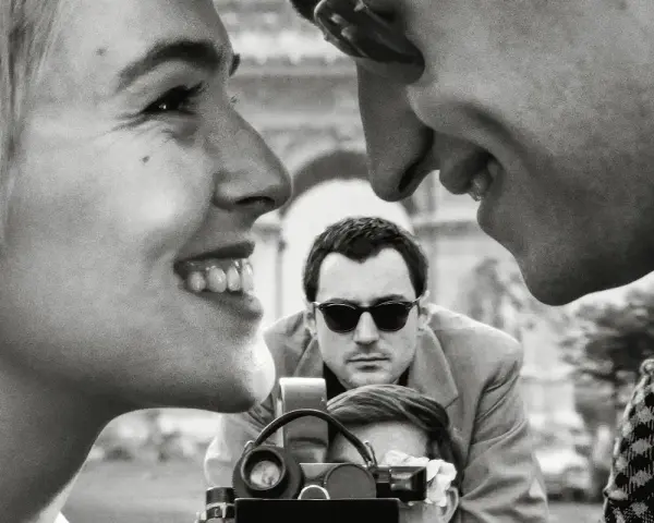 Primo piano di una coppia che ride, con un uomo che filma sullo sfondo, evocando l'estetica dei registi della nouvelle vague.