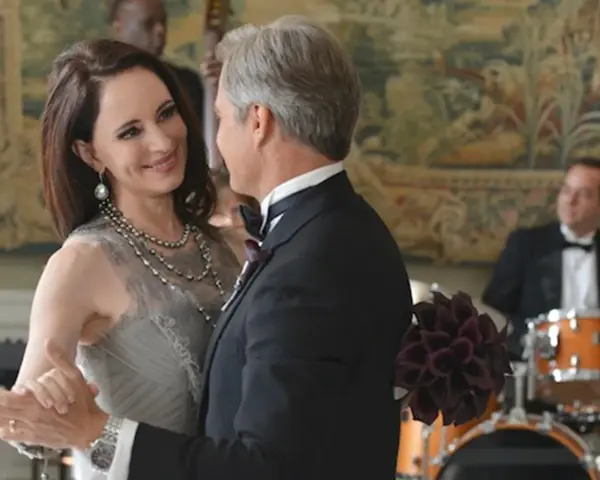 Il cast di Revenge balla elegantemente. Una donna con un abito grigio e collane di perle, un uomo in smoking.