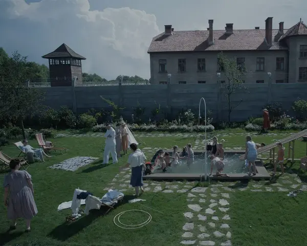 Scena da film strani: bambini giocano in piscina, adulti sorvegliano, mentre sullo sfondo incombono torri di guardia e un edificio imponente.