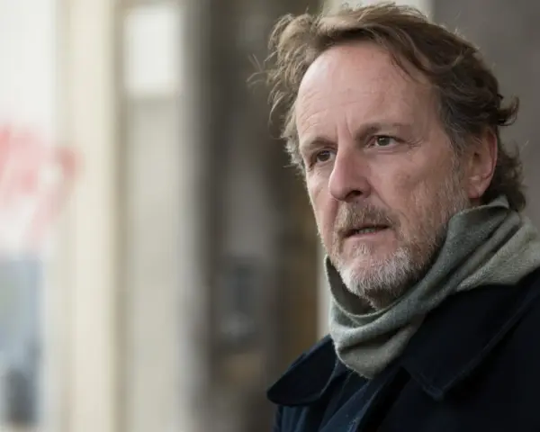 Christian Peppard, con un foulard e un cappotto scuro, guarda pensieroso verso destra.