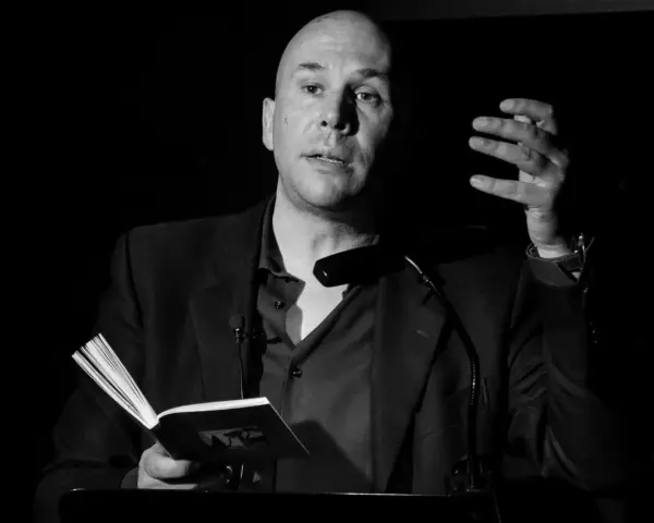 Un uomo calvo, narratore protagonista, legge da un libro aperto, gesticolando con la mano.