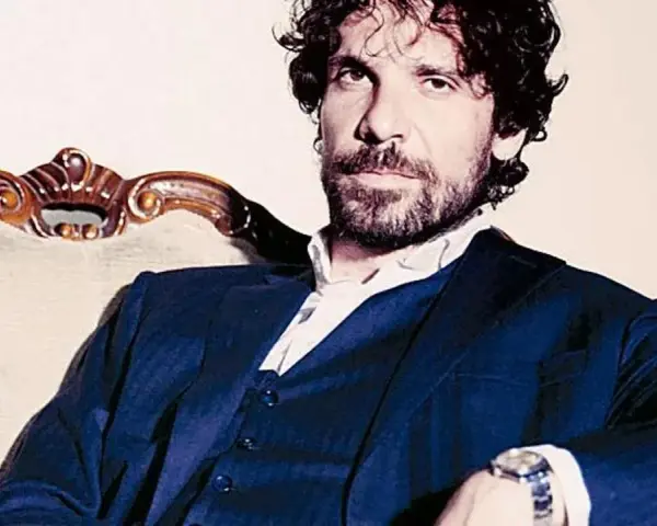 L'attore Montanari, con barba e capelli ricci, indossa un completo blu e una camicia bianca, seduto su una poltrona d'epoca.