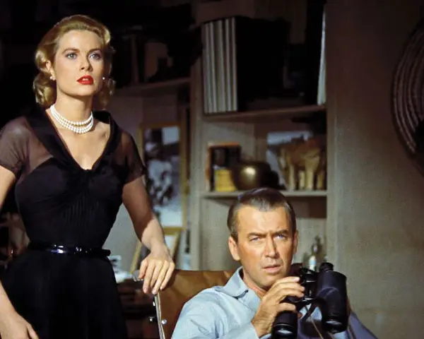 Grace Kelly e James Stewart in una scena di un film thriller suspense. Lui osserva con il binocolo, lei elegante in abito nero.