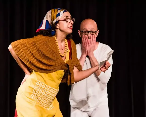 Due attori in scena, una donna con foulard e grembiule giallo, un uomo con le mani sul volto. Una scena di commedia drammatica.