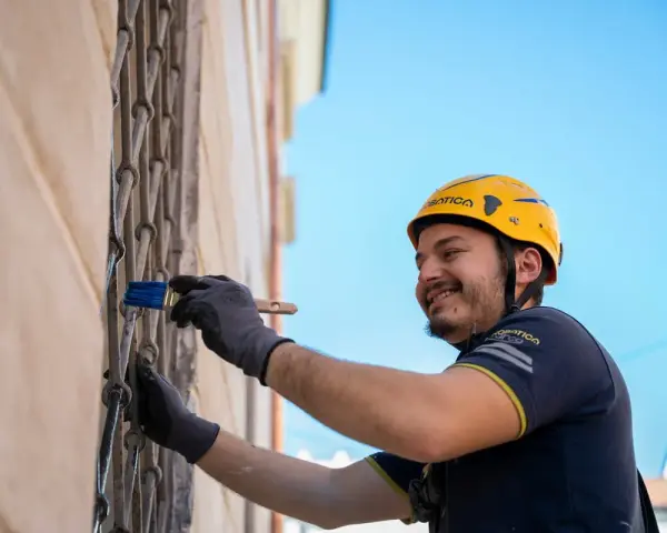 Uomo con casco giallo dipinge una grata metallica con un pennello blu. Inquadratura dal basso, cielo azzurro sullo sfondo.