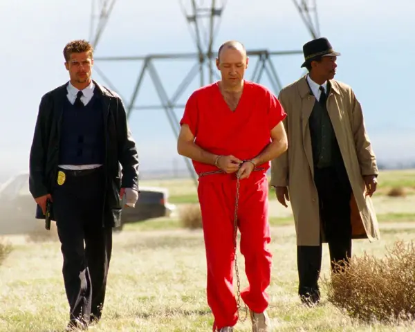 Tre uomini camminano in un campo: un detenuto in tuta rossa ammanettato, un poliziotto e un detective. Scena da film polizieschi recenti.