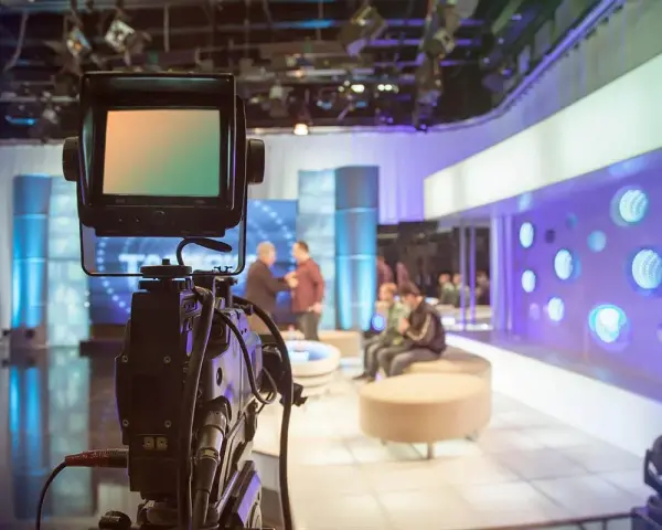 Telecamera in primo piano in uno studio televisivo. Si intravede il backstage, con persone sedute e in piedi, luci soffuse e un'atmosfera di attesa.