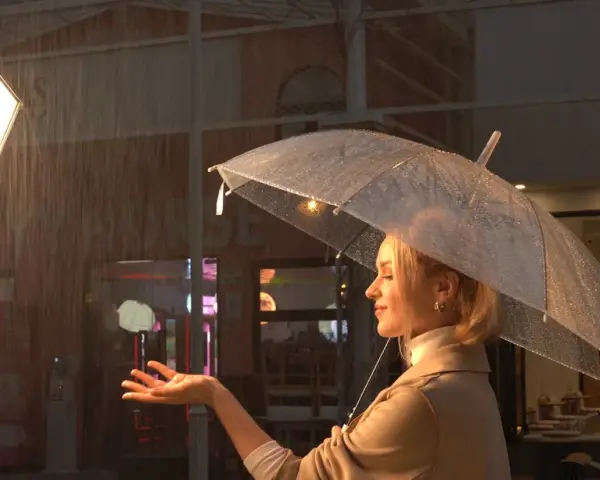 Una donna con ombrello trasparente sotto la pioggia, illuminata da potenti luci set cinematografico. La scena evoca un'atmosfera suggestiva e cinematografica.