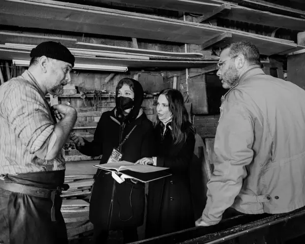 Foto di scena: un artigiano discute con due donne, una con mascherina, mentre un uomo osserva.