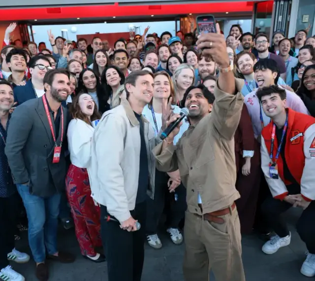 Un gruppo di persone sorride e fa un selfie a Hollywood, America. Alcuni tengono microfoni, creando un'atmosfera di festa.