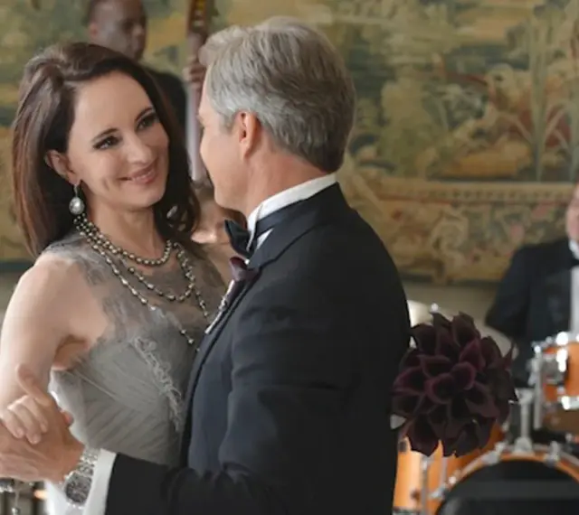 Il cast di Revenge balla elegantemente. Una donna con un abito grigio e collane di perle, un uomo in smoking.