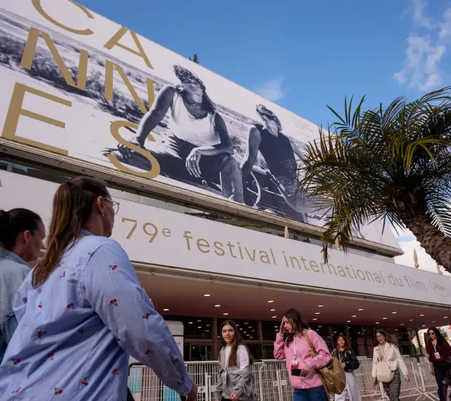 Persone camminano sotto il cartellone del Festival di Cannes, un evento che celebra il cinema internazionale, rivaleggiando con l'atmosfera di Hollywood.